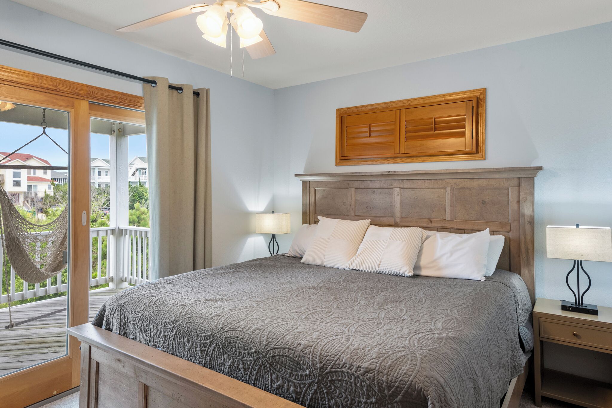 A bedroom with a bed, a wooden headboard, two bedside lamps, a ceiling fan, and a view of a balcony.
