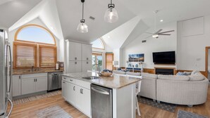 Una cocina moderna con armarios blancos, una isla central, electrodomésticos de acero inoxidable y un amplio ventanal con persianas de madera.