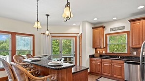A kitchen with wooden cabinets, a central island with seating, and a view of the outdoors through large windows. - Sea-Esta: Private Pool | Hot Tub | Foosball Table (Corolla)