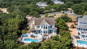 A large, two-story house with a swimming pool and multiple decks, surrounded by dense trees and a view of a lake. - Sea-Esta: Private Pool | Hot Tub | Foosball Table (Corolla)