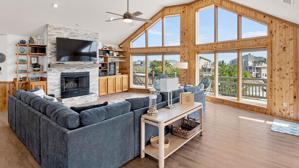 A spacious living room with a large gray sectional sofa, a fireplace, wooden shelves, a flat-screen TV, and large windows offering a view of the neighborhood. - The Wheelhouse: Pool and Hot Tub | 500ft. to Beach (Corolla)