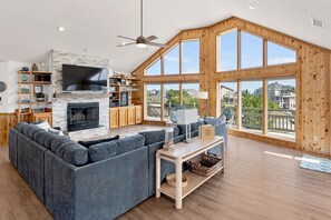 A spacious living room with a large gray sectional sofa, a fireplace, wooden shelves, a flat-screen TV, and large windows offering a view of the neighborhood. - The Wheelhouse: Pool and Hot Tub | 500ft. to Beach (Corolla)