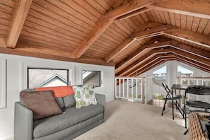 Ein geräumiges Dachzimmer mit hoher Holzbalkendecke, einem grauen Sofa mit gemusterten Kissen, einem Esstisch mit Stühlen und großen Fenstern mit Blick auf die Dachlinie.
