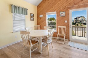A dining area with a round table and six chairs, wooden walls, and a view of a residential area through a glass door. - Crabby Patty: Pool Access | Short Walk to Beach (Nags Head)