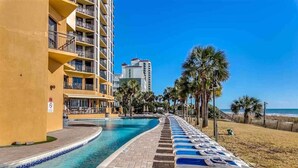 A pool area with lounge chairs, a building with balconies, and a beach in the background. - 1510 Pearl on the Beach. Pet Friendly. Oceanfront. (Myrtle Beach)