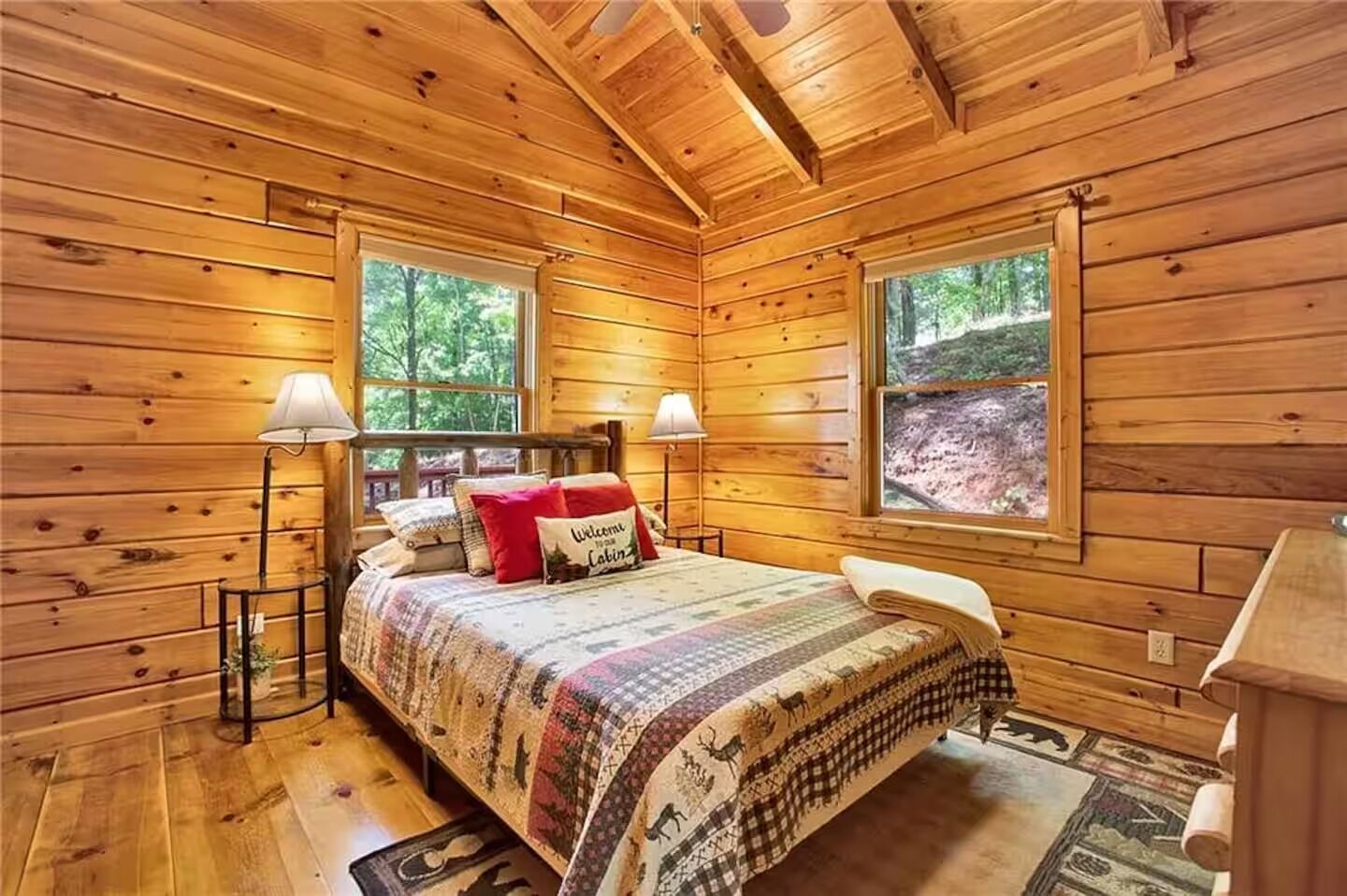 A wooden cabin bedroom with a bed, two bedside lamps, a window, and a welcome sign.