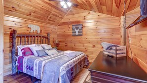 Un dormitorio de cabaña de madera con una cama, un ventilador de techo, un televisor montado y una estantería en la pared.