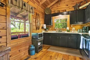 A wooden cabin interior with a kitchen area, including cabinets, a stove, and a window overlooking greenery.
