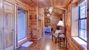 A wooden cabin interior with a hallway leading to a bedroom, a dining area with a table and chairs, and a large window providing natural light.