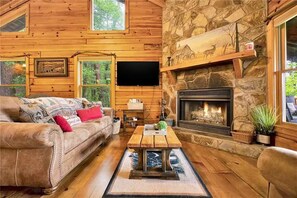 A cozy cabin interior with a stone fireplace, wooden walls, a leather sofa, and a flat-screen TV.