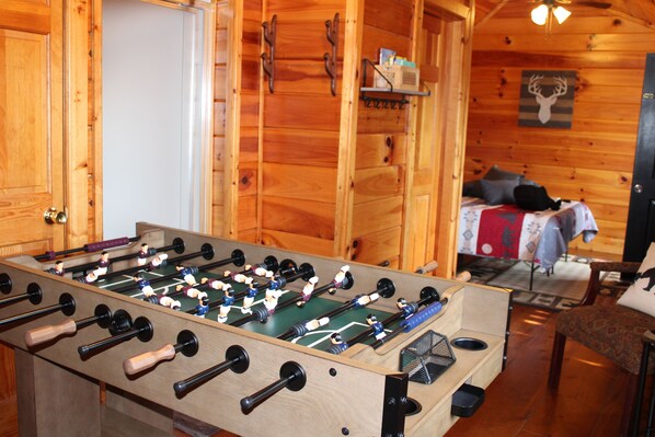 A wooden-floored room with a foosball table, a bed with pillows, and a shelf with items.