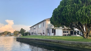 Ein zweistöckiges Haus mit einer überdachten Terrasse, einem großen Baum davor und einem Kanal daneben.