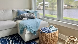 Ein Wohnzimmer mit einer Couch, Kissen, einer blauen Decke, einem gewebten Korb und Blick auf einen See mit Häusern im Hintergrund.