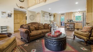 A spacious living room with a high ceiling, a large sofa, a coffee table, and a dining area with wooden cabinets and a kitchen in the background. - Cozy Cottage minutes from Wineries & 5 miles to Dahlonega Square (Dahlonega)