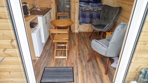 A compact room with a wooden floor, a small kitchenette, a round wooden table with chairs, a grey armchair, and a bed with a plaid blanket. - Anneth Glamping Pod (Sleat)