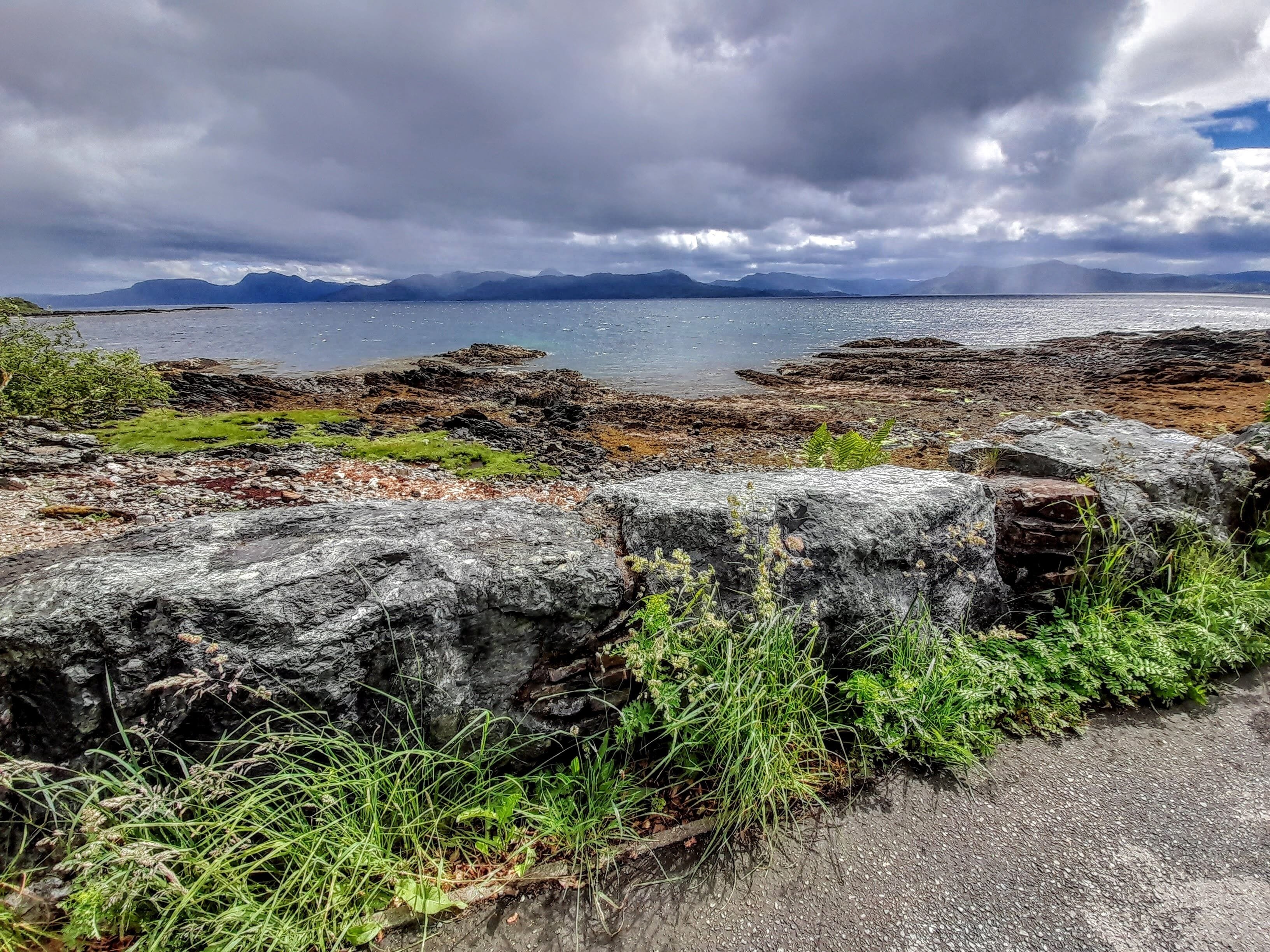 A coastal landscape with rocky terrain, a body of water, and distant mountains under a cloudy sky.