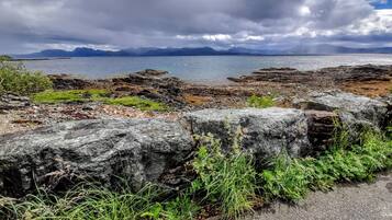 A coastal landscape with rocky terrain, a body of water, and distant mountains under a cloudy sky.