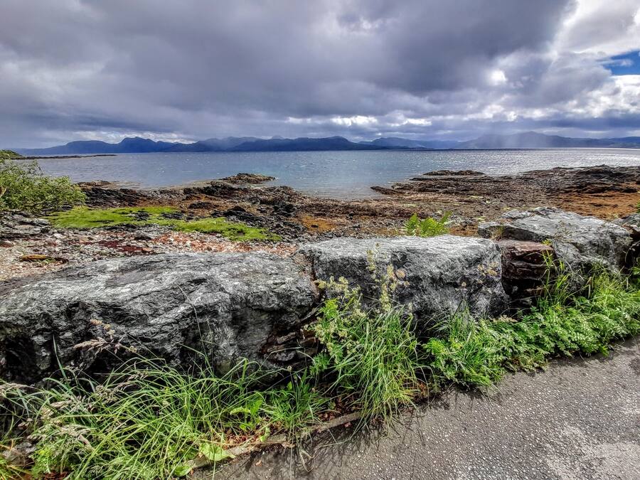 A coastal landscape with rocky terrain, a body of water, and distant mountains under a cloudy sky.