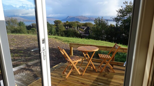 A wooden deck with a table and chairs, overlooking a grassy area and a body of water with mountains in the distance. - Anneth Glamping Pod (Sleat)