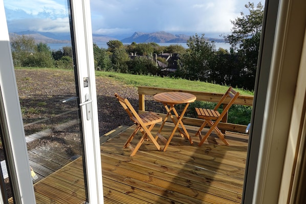 Une terrasse en bois avec une table et des chaises, donnant sur une pelouse et un plan d’eau, avec des montagnes visibles au loin.