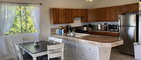 Una cocina con armarios de madera, un refrigerador de acero inoxidable, una encimera de mármol y un comedor con sillas blancas y una mesa.
