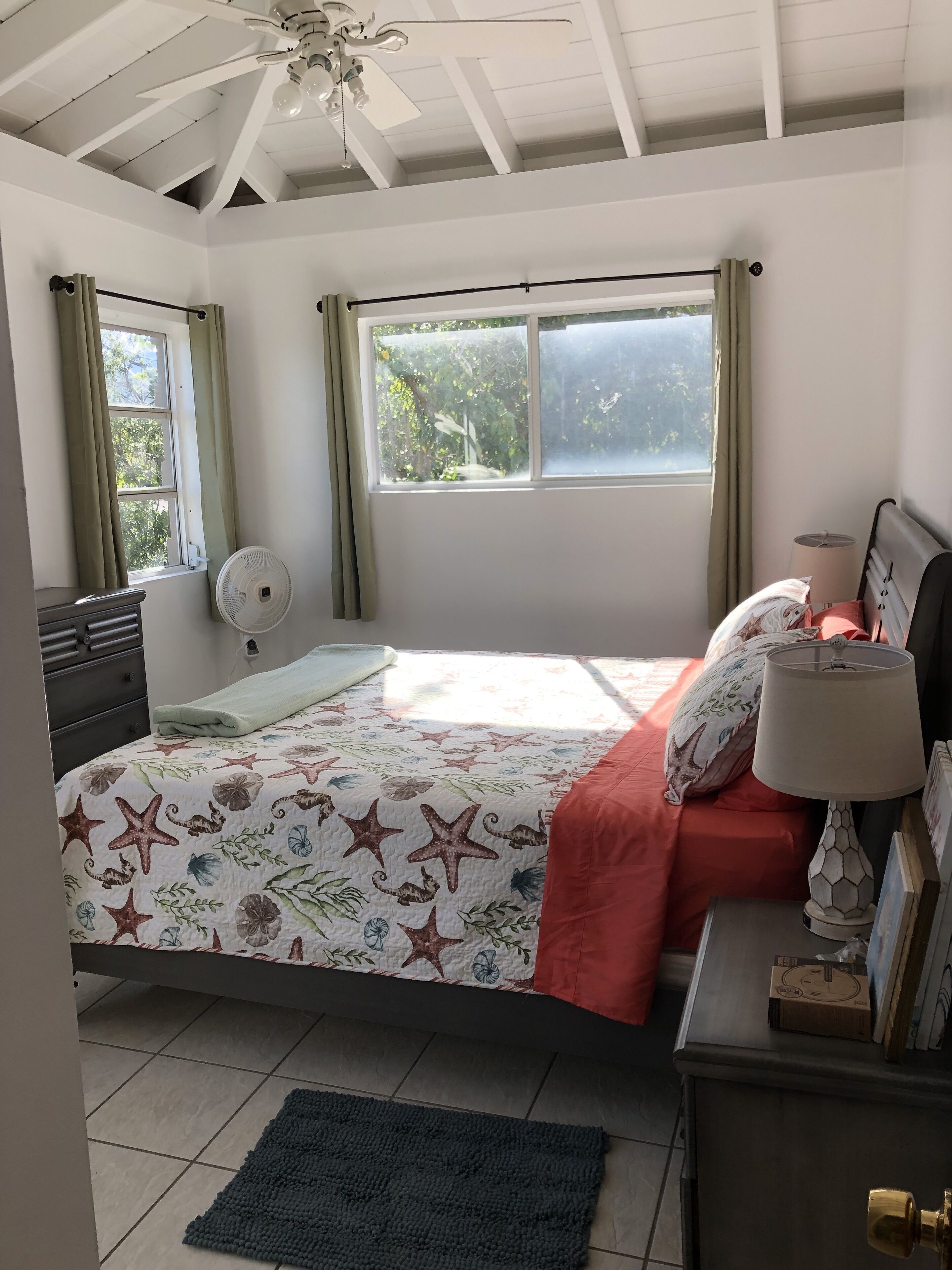 A bedroom with a bed, a nightstand, a desk, a fan, and a window with curtains.