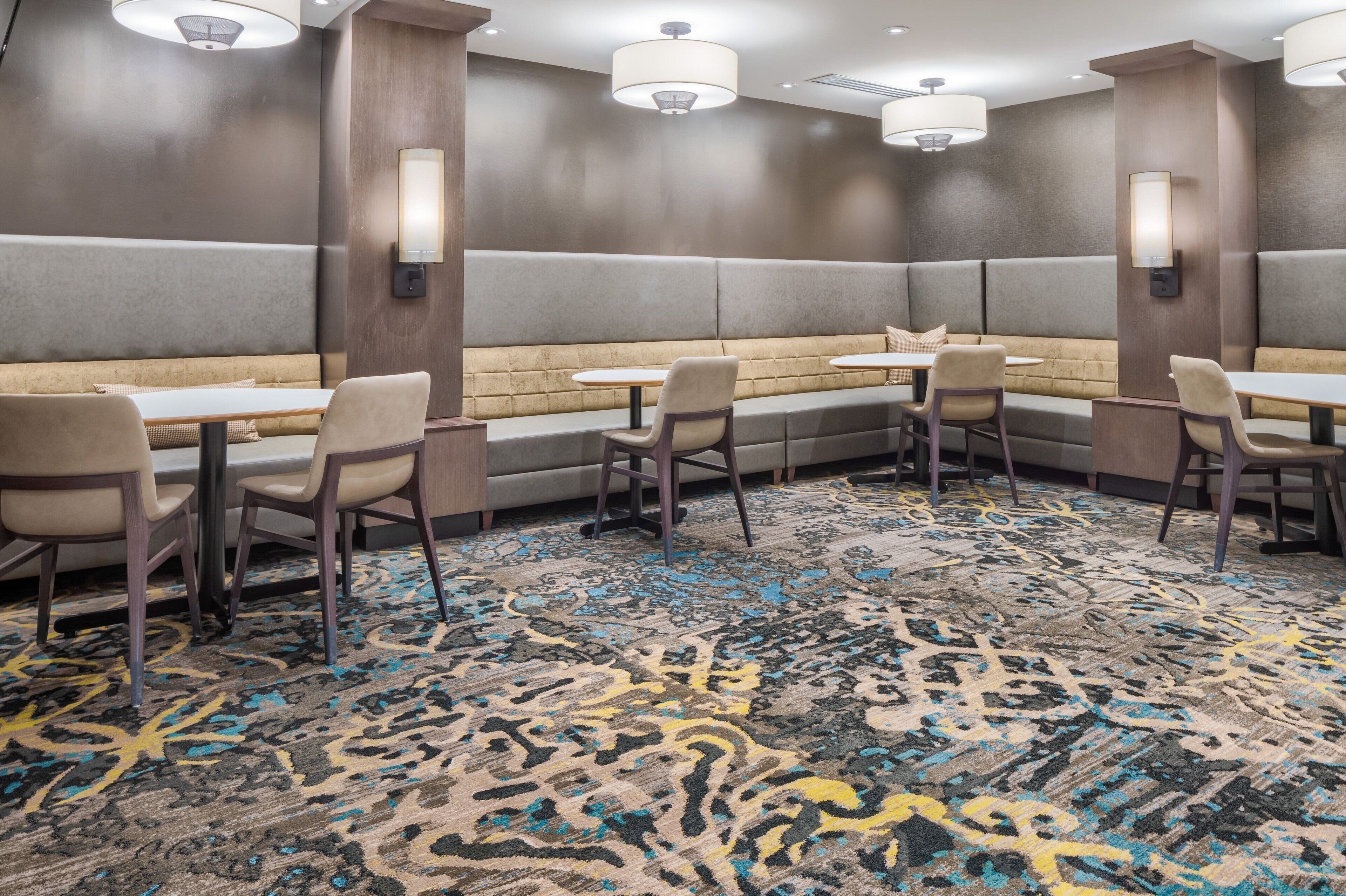 A hotel room with a seating area featuring cushioned benches and tables, a patterned carpet, and wall-mounted lighting.