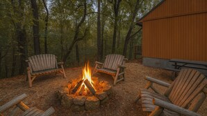 A campfire with four Adirondack chairs arranged around it, set in a forested area with a wooden building in the background.