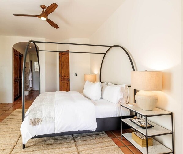 A bedroom with a four-poster bed, a ceiling fan, a bedside table with a lamp, and a nightstand with a tray.