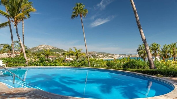 A swimming pool with a stone deck, surrounded by palm trees and a view of a distant town and hills. - Peaceful Palmilla Ocean View Oasis Steps to Beach (Palmilla)