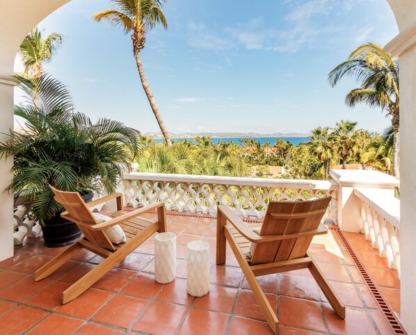A balcony with two wooden chairs and a white table, overlooking a coastal landscape with palm trees and a view of the sea.