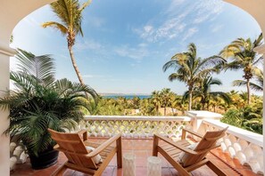 A terrace with wooden chairs and a potted plant, overlooking a coastal landscape with palm trees and a view of the sea.