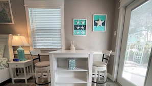 Una habitación con un escritorio blanco, dos sillas, una mesita con una lámpara y una ventana con persianas.