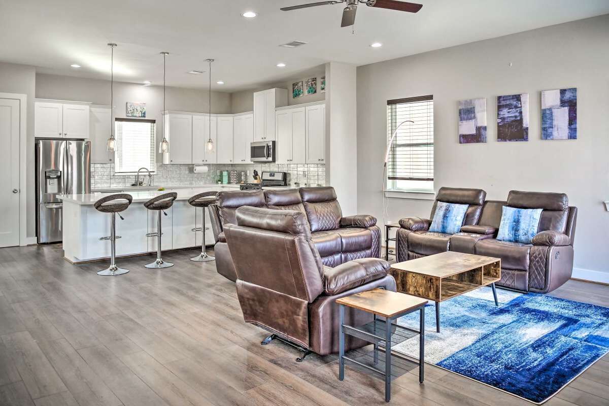 Un salon moderne avec un fauteuil inclinable en cuir et une table basse, une cuisine avec des meubles blancs et des appareils en acier inoxydable, et un coin repas avec des tabourets de bar.
