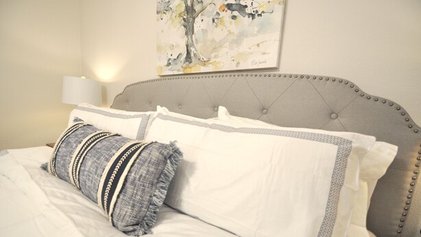 Una cama bien hecha con ropa de cama blanca y gris, un cabecero tapizado en gris con adornos de borlas, y una lámpara fijada a la pared.
