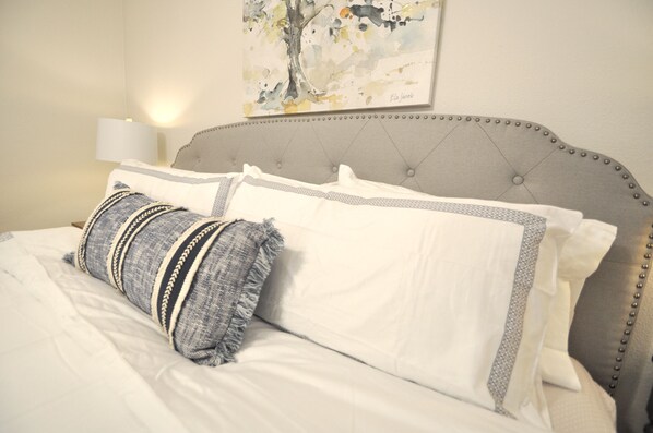 A neatly made bed with white and gray bedding, a gray upholstered headboard with decorative bead trim, and a wall-mounted lamp.