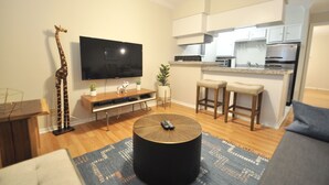 Un moderno salón con un televisor de pantalla plana, una escultura de jirafa, una mesa de centro redonda y una zona de cocina con taburetes de bar.