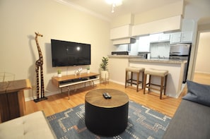 A modern living room with a flat-screen TV, a giraffe sculpture, a round coffee table, and a kitchen area with bar stools.