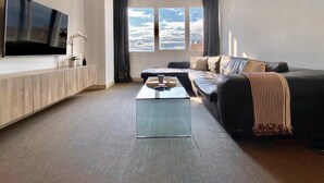 A modern living room with a large flat-screen TV, a leather sofa, a coffee table, and a view of the sky through large windows. - Charming Eurobuilding Skyline Madrid - Spectacular Views (Madrid)