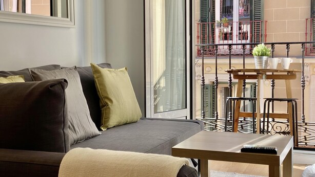 Ein modernes Wohnzimmer mit einem grauen Sofa, einem Holztisch und Blick auf einen Balkon mit Geländer und Blumentöpfen.