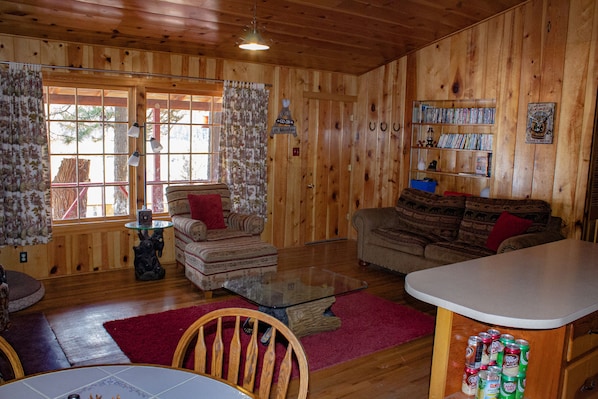 Un interior acogedor de cabaña con paredes de madera, una mesa de centro de vidrio, dos sofás, una mesa de comedor y una estantería llena de libros y DVD.