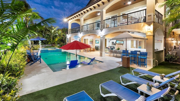 A two-story building with a swimming pool, outdoor seating, and a well-maintained garden. - Elev • 5BR/8BA • Game Room • 75’ Deep Canal Dock • Ice Maker • Kayaks • Bikes (Key Colony Beach)