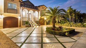 A two-story house with a well-lit entrance, a staircase, and a landscaped garden with palm trees. - Elev • 5BR/8BA • Game Room • 75’ Deep Canal Dock • Ice Maker • Kayaks • Bikes (Key Colony Beach)