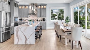 A modern kitchen with grey cabinets, a marble island, and a dining area with white chairs. - Elev • 5BR/8BA • Game Room • 75’ Deep Canal Dock • Ice Maker • Kayaks • Bikes (Key Colony Beach)