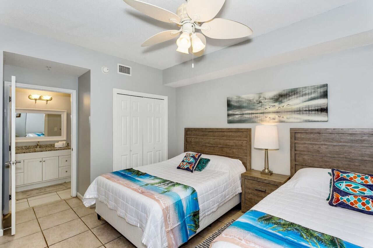 A hotel room with two beds, a ceiling fan, a painting, and a bathroom with a sink and mirror.