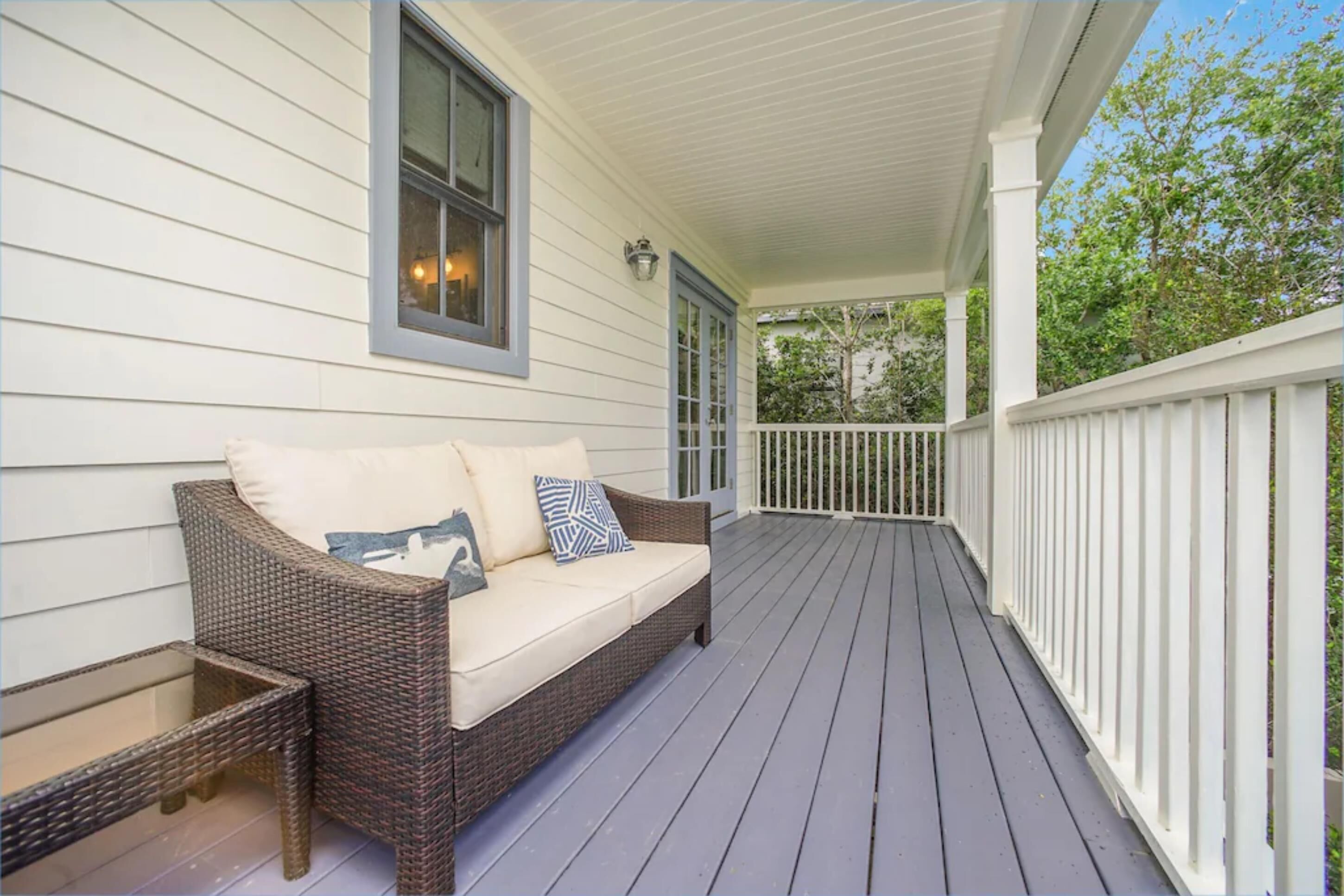 A covered porch with a wicker sofa and a glass table, white railings, and a view of trees.