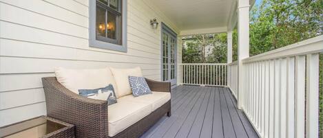 A covered porch with a wicker sofa and a glass table, white railings, and a view of trees.