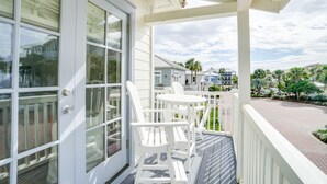 A covered porch with white chairs and a table, a white door with glass panels, and a view of a street with buildings and palm trees. - 30A Beach House - Stillwater Cottage By Panhandle Getaways (Panama City Beach)