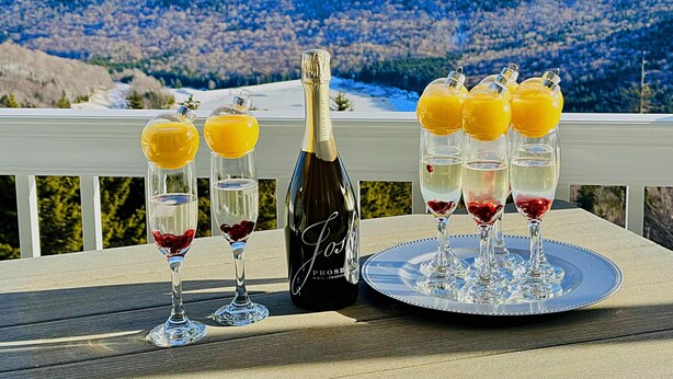 A table with a bottle of wine and glasses of red wine and orange juice on a deck with a scenic mountain view in the background. - ML 248: Your unique mountain biking haven with incredible views! (Snowshoe)