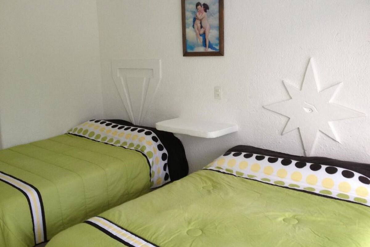 Una habitación con dos camas, cada una con ropa de cama verde y almohadas de lunares. Se observa un estante blanco y un adorno en la pared con forma de estrella.